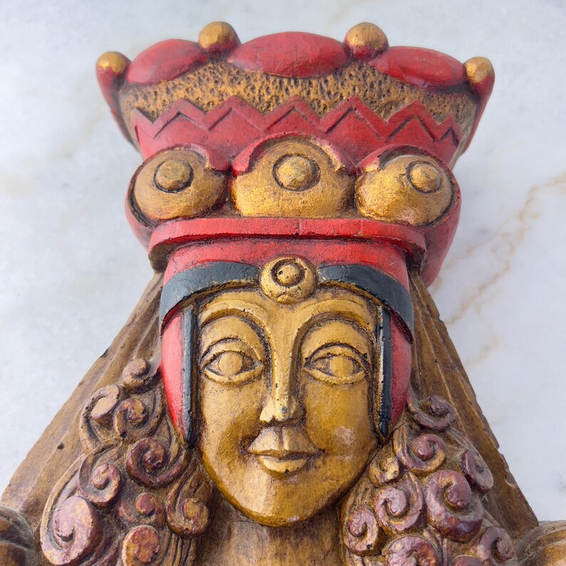 May include: A wooden sculpture of a woman's face with a red and gold crown. The woman's face is painted gold and has a serene expression. The crown is decorated with gold circles and has a red base.