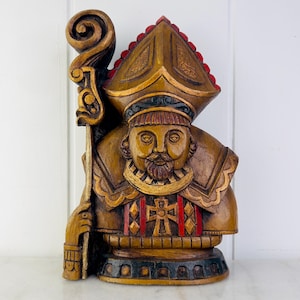 May include: A wooden sculpture of a bishop wearing a red and gold mitre and holding a staff. The sculpture is painted in shades of brown, gold, and red.