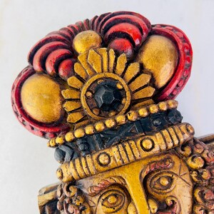 May include: A wooden wall sculpture of a face with a gold and red crown. The face has a detailed design with a black jewel in the centre of the crown.