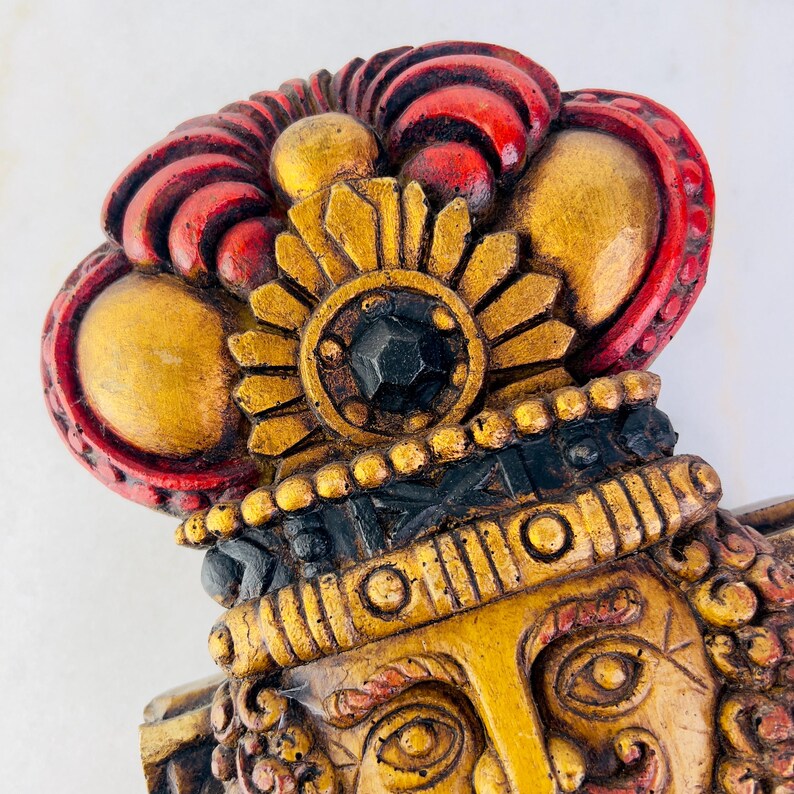 May include: A wooden wall sculpture of a face with a gold and red crown. The face has a detailed design with a black jewel in the centre of the crown.