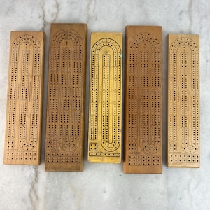 Op de afbeelding: Vijf houten cribbageborden in verschillende bruin- en geeltinten. Elk bord heeft een reeks gaten om te scoren, met een gebogen bovenkant en een rechthoekige vorm. De borden zijn voor het klassieke kaartspel.