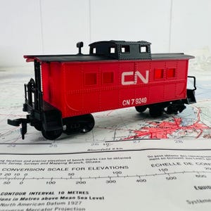 May include: A red and black toy train caboose with the text "CN 7 9249" in white. The caboose has a black roof and wheels. The toy is sitting on a map.