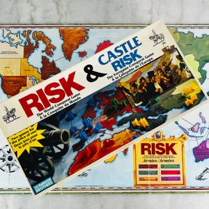 May include: A Risk & Castle Risk board game box featuring a map of Europe, a black cannon illustration, and the text "Risk & Castle Risk The World Conquest Game". The box includes yellow and blue plastic army pieces and a Parker Brothers logo.