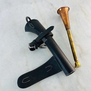 May include: A black leather hunting horn case with white stitching and a buckle closure. Beside it is a brass and copper hunting horn, featuring a flared copper bell and a brass body.