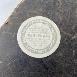 May include: A white round sticker with black text that reads "Fragile - Hang Indoors Only". The sticker has a seal that certifies that the work of art is cast entirely of solid-core Vanathane. The text also reads "Vanathane" and "Used only in fine sculpture where pride in craft is a cherished tradition."
