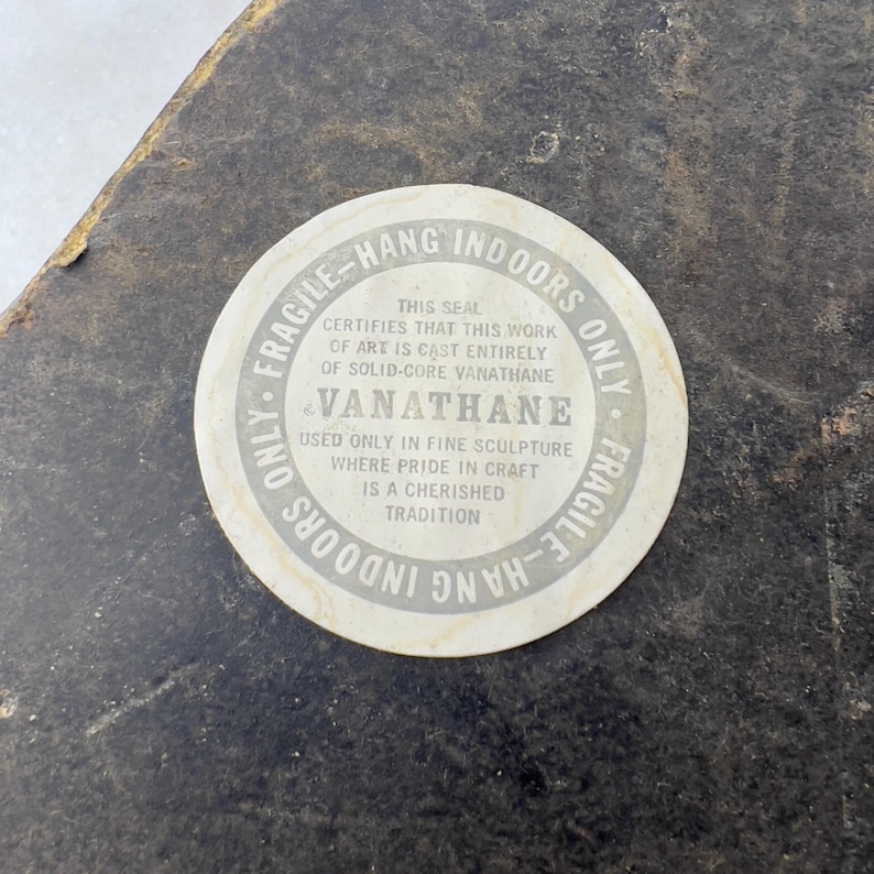 May include: A white round sticker with black text that reads "Fragile - Hang Indoors Only". The sticker has a seal that certifies that the work of art is cast entirely of solid-core Vanathane. The text also reads "Vanathane" and "Used only in fine sculpture where pride in craft is a cherished tradition."