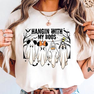 Può includere: T-shirt color crema con la scritta "HANGIN WITH MY BOOS" sopra fantasmi dei cartoni animati che assomigliano a Topolino, Minnie, Paperino e Pippo. I fantasmi sono appesi a una linea nera con pipistrelli e alberi. La maglietta è abbinata a pantaloncini di jeans blu.