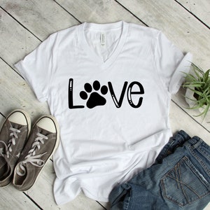 May include: White V-neck t-shirt with the word "LOVE" in black lettering with a paw print in the center of the "O". The shirt is laying on a white wood surface with a pair of gray sneakers and a pair of blue denim shorts.