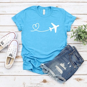 May include: A turquoise blue t-shirt with a white airplane and heart design. The airplane is flying away from a heart.