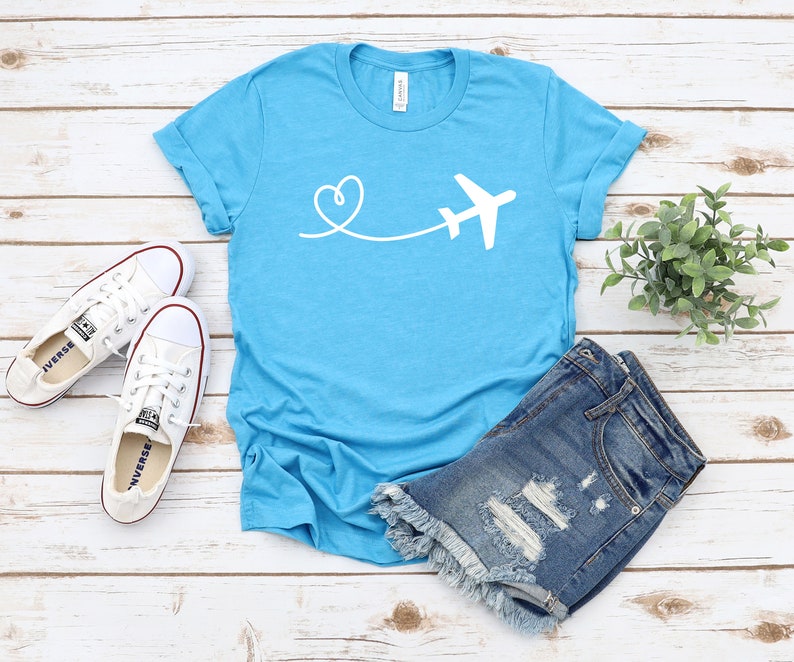 May include: A turquoise blue t-shirt with a white airplane and heart design. The airplane is flying away from a heart.