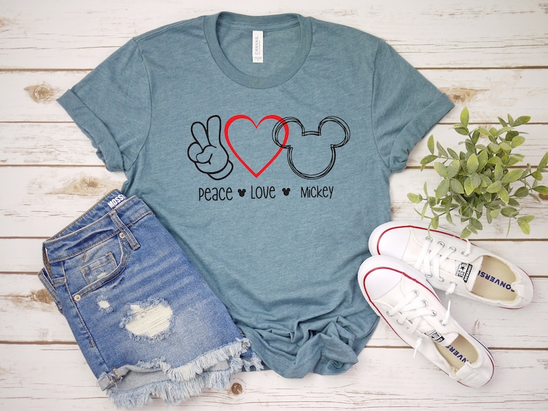 Puede incluir: Una camiseta azul turquesa con un signo de paz, un coraz&oacute;n y un gr&aacute;fico de la cabeza de Mickey Mouse. El texto "Peace Love Mickey" est&aacute; impreso debajo del gr&aacute;fico.