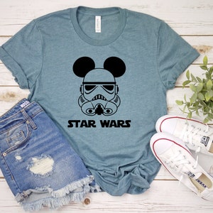 May include: A blue t-shirt featuring a black graphic of a Stormtrooper helmet with Mickey Mouse ears and the words "STAR WARS" in black. The shirt is paired with denim shorts and white sneakers.
