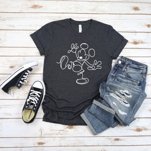 Puede incluir: Una camiseta gris oscuro con un contorno blanco de Mickey Mouse en un estilo de dibujo lineal simple. La camiseta est&aacute; colocada sobre un fondo de madera blanca con un par de zapatillas Converse negras y un par de pantalones vaqueros azules.