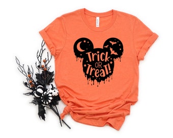 Puede incluir: Una camiseta naranja de manga corta con un gráfico negro de una silueta de orejas de ratón. El diseño incluye una luna creciente, murciélagos, estrellas y el texto Trick or Treat! en naranja con un efecto de goteo en el borde inferior.