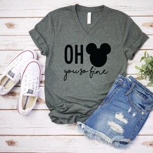 Może przedstawiać: Zielony T-shirt z dekoltem w kształcie litery V z napisem "OH you so fine" i czarnym nadrukiem głowy Myszki Miki. Koszulka jest połączona z przetartymi dżinsowymi szortami i białymi butami sportowymi.