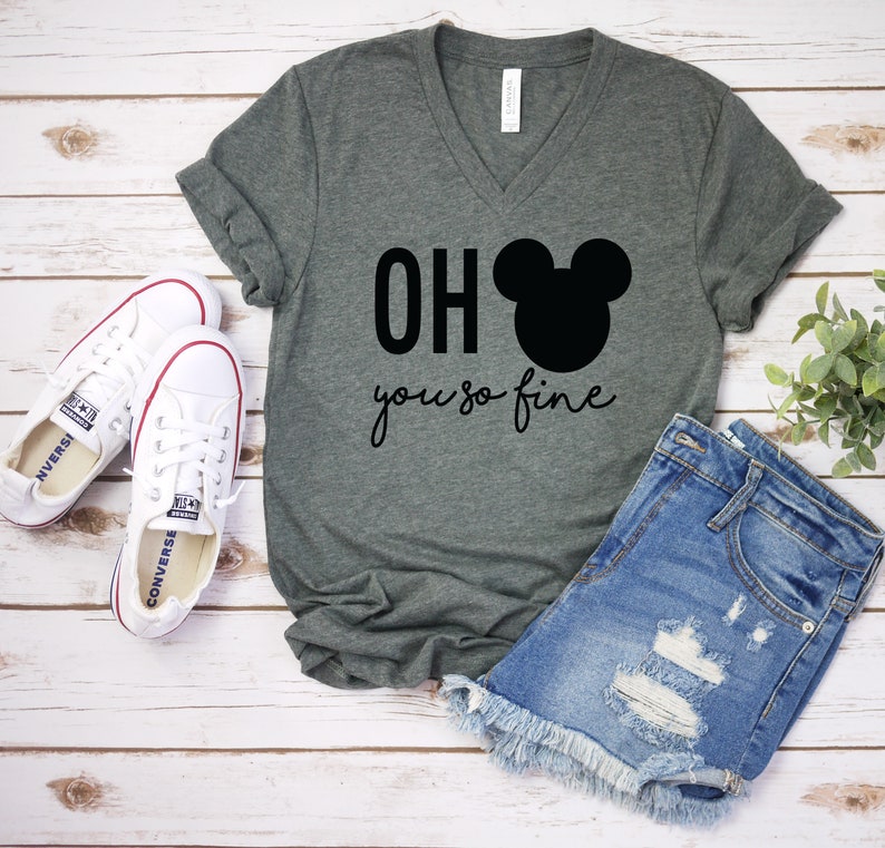 Może przedstawiać: Zielony T-shirt z dekoltem w kształcie litery V z napisem "OH you so fine" i czarnym nadrukiem głowy Myszki Miki. Koszulka jest połączona z przetartymi dżinsowymi szortami i białymi butami sportowymi.