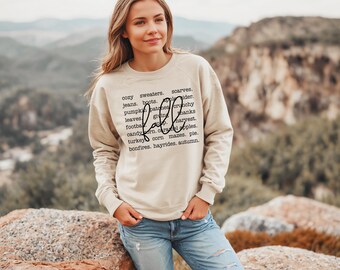May include: A beige sweatshirt with the word "fall" in black script surrounded by a list of fall-related words, such as "cozy sweaters", "scarves", "jeans", "boots", "pumpkin patches", "crunchy leaves", "giving thanks", "football", "harvest", "candy corn", "caramel apples", "turkey", "corn mazes", "pie", "bonfires", "hayrides", and "autumn".