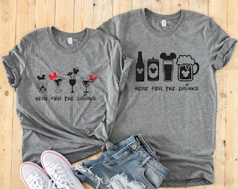 May include: Two gray t-shirts with a black and white graphic design of Mickey Mouse ears and drinks. The left shirt says "Here for the drinks" and features a beer mug, a martini glass, a wine glass, and a cocktail glass. The right shirt says "Here for the drinks" and features a beer bottle, a beer can, a beer glass, and a beer mug.
