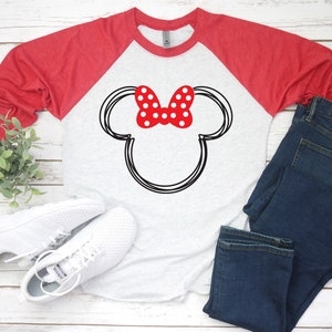 May include: A white and red baseball tee shirt with a black and white outline of Mickey Mouse's ears and a red bow with white polka dots. The shirt is a raglan style with red sleeves and a white body. The shirt is lying on a white wooden surface with a pair of white trainers and blue jeans.
