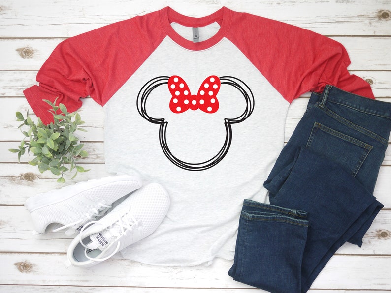 May include: A white and red baseball tee shirt with a black and white outline of Mickey Mouse's ears and a red bow with white polka dots. The shirt is a raglan style with red sleeves and a white body. The shirt is lying on a white wooden surface with a pair of white trainers and blue jeans.