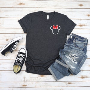 K&ouml;nnte beinhalten: Ein dunkelgraues T-Shirt mit einer wei&szlig;en Umrandung von Mickey Mouse' Kopf mit einer roten Schleife oben. Das Shirt liegt auf einer wei&szlig;en Holzoberfl&auml;che mit einem Paar schwarzen Converse-Turnschuhen und einer blauen Jeans.