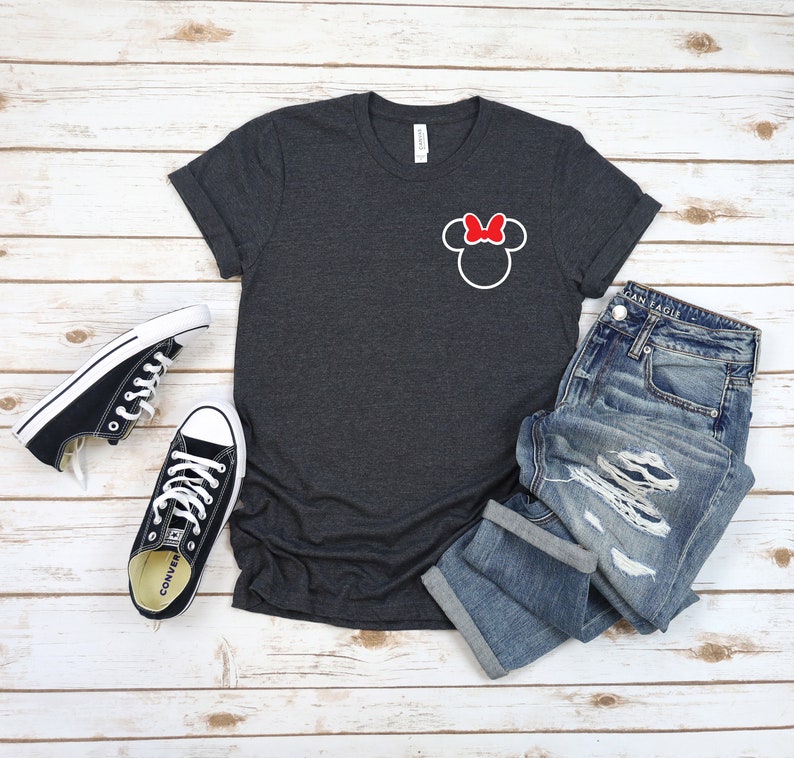 K&ouml;nnte beinhalten: Ein dunkelgraues T-Shirt mit einer wei&szlig;en Umrandung von Mickey Mouse' Kopf mit einer roten Schleife oben. Das Shirt liegt auf einer wei&szlig;en Holzoberfl&auml;che mit einem Paar schwarzen Converse-Turnschuhen und einer blauen Jeans.