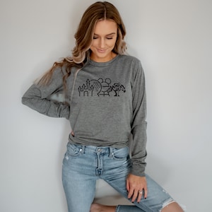 Puede incluir: Una camiseta de manga larga gris jaspeada con un gráfico negro de un castillo, una esfera, una montaña rusa y un árbol. El gráfico es una representación estilizada de un famoso parque de atracciones.