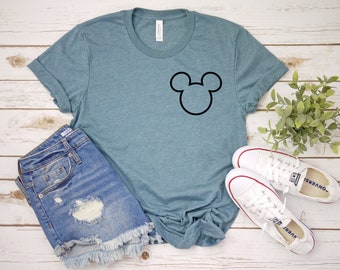 May include: A teal blue t-shirt with a black Mickey Mouse head silhouette on the front. The t-shirt is paired with denim shorts and white sneakers.