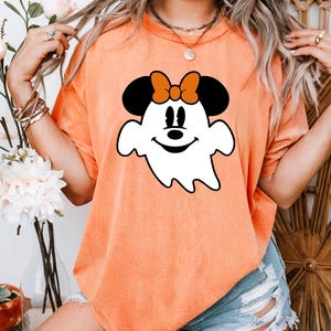 Può includere: T-shirt arancione con un design a forma di fantasma dei cartoni animati, con orecchie e fiocco di Minnie Mouse. Il fantasma ha una faccia sorridente ed è delineato in nero. La maglietta è di colore arancione.