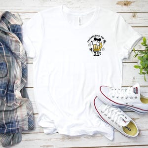 May include: White t-shirt featuring a beer mug graphic with Mickey Mouse ears and the text "Celebrating My 21st". The shirt is styled with white trainers with red and blue details and a plaid shirt.
