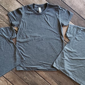 May include: Three plain gray t-shirts of different sizes, laid out on a woodgrain surface.