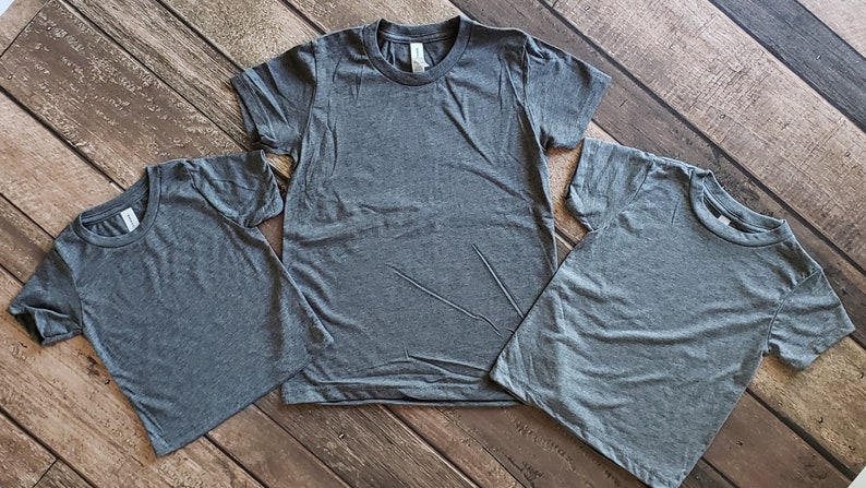 May include: Three plain gray t-shirts of different sizes, laid out on a woodgrain surface.