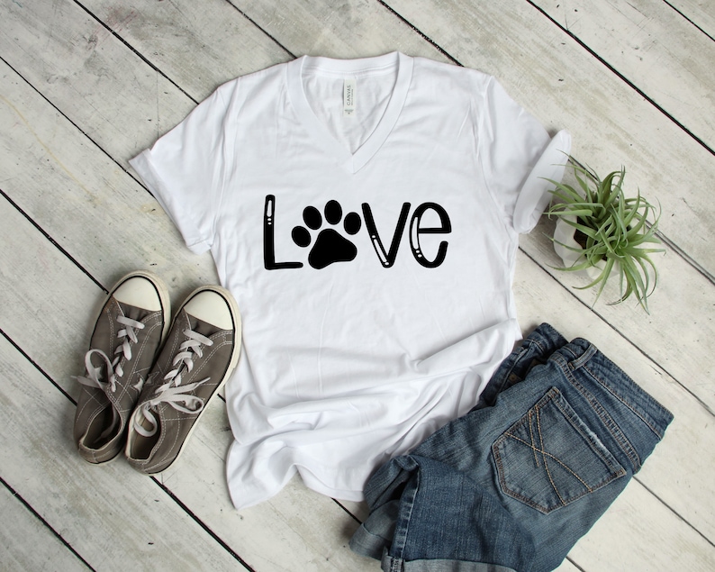 May include: White V-neck t-shirt with the word "LOVE" in black lettering with a paw print in the center of the "O". The shirt is laying on a white wood surface with a pair of gray sneakers and a pair of blue denim shorts.