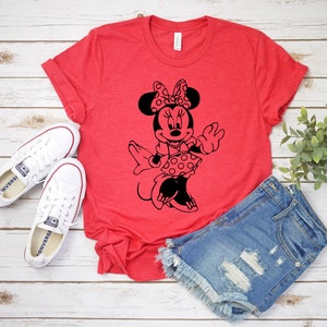 May include: Red t-shirt with a black and white graphic of Minnie Mouse wearing a polka dot dress and bow. The shirt is on a white wooden surface with a pair of white sneakers and denim shorts.