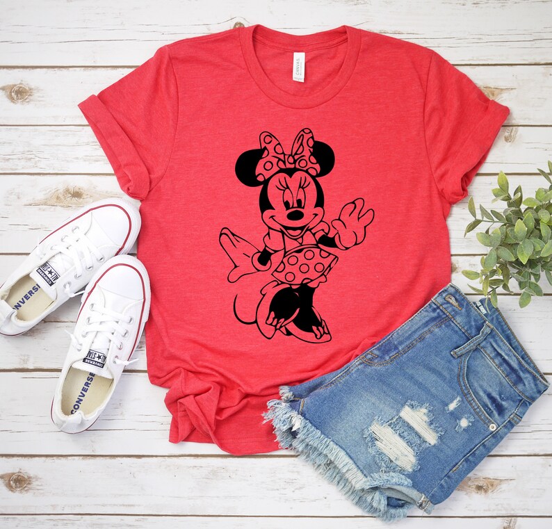 May include: Red t-shirt with a black and white graphic of Minnie Mouse wearing a polka dot dress and bow. The shirt is on a white wooden surface with a pair of white sneakers and denim shorts.