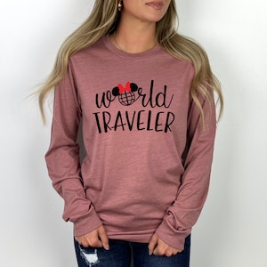 Puede incluir: Una camiseta de manga larga de color malva con un gráfico negro que dice "world traveler" y una cabeza de Mickey Mouse roja y negra con un globo en el medio.