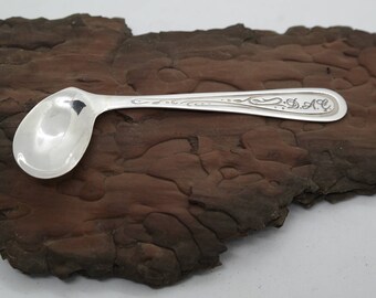 May include: A silver teaspoon with an ornate handle and the initials "S.A.C." engraved on the back. The spoon is resting on a piece of brown wood.