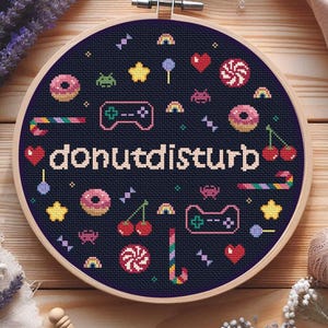 Op de afbeelding: Een kruissteek borduurring met een marineblauwe achtergrond. Het ontwerp toont de tekst "donutdisturb" omringd door kleurrijke afbeeldingen van donuts, harten, sterren en gamecontrollers. De ring is geplaatst in een houten frame.