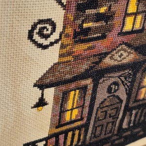 The image is a close-up of a cross-stitch embroidery depicting a house with a porch, featuring a pumpkin and a spider web.