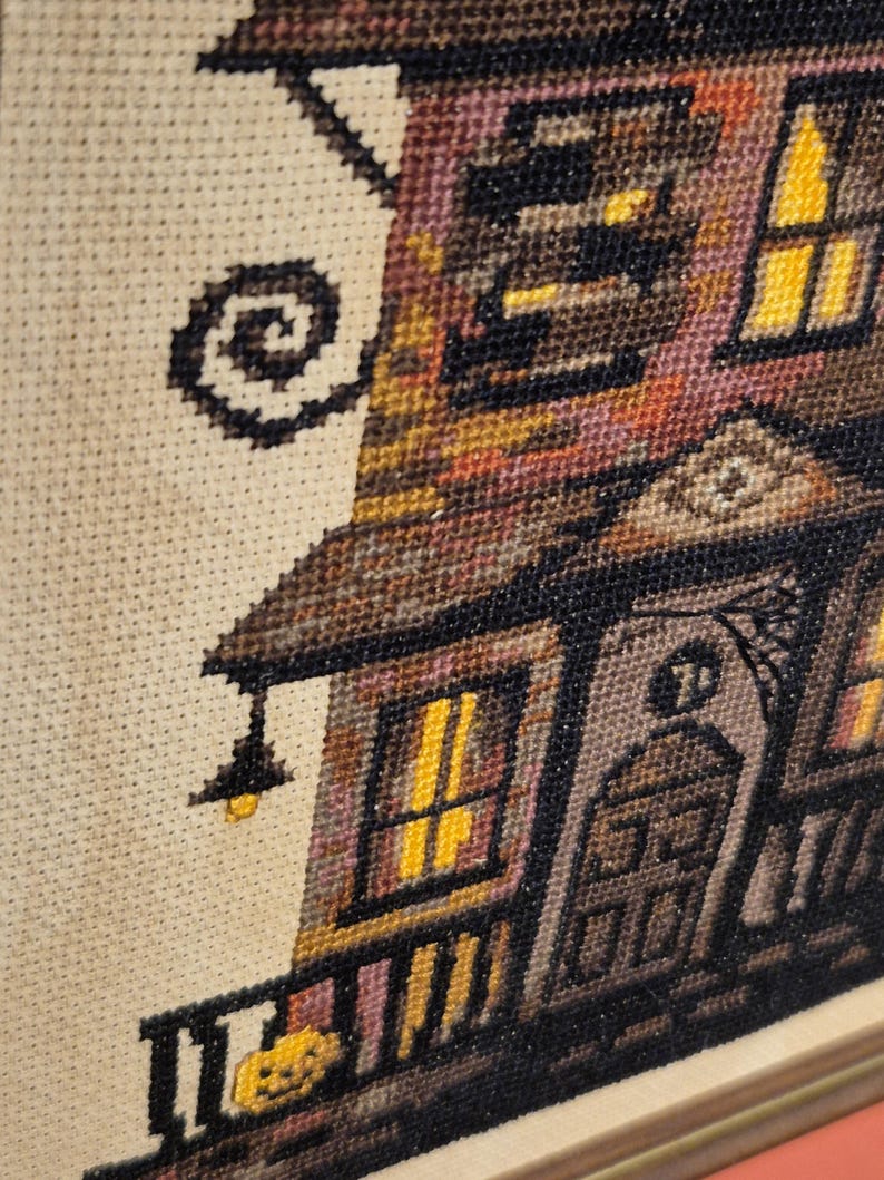 The image is a close-up of a cross-stitch embroidery depicting a house with a porch, featuring a pumpkin and a spider web.