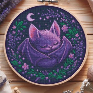 Könnte beinhalten: Ein Kreuzstich-Stickrahmen mit einer schlafenden lila Fledermaus mit ausgebreiteten Flügeln, einem Halbmond und Sternen auf einem dunkelblauen Hintergrund. Die Fledermaus ist von einem floralen Kranz aus Lavendel und rosa Blüten umgeben, alles in einem Holzrahmen.