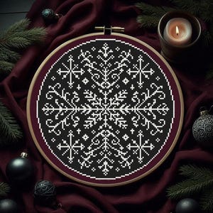 Könnte beinhalten: Ein schwarz-weißes Kreuzstich-Schneeflocken-Design in einem hölzernen Stickrahmen. Das Design ist umgeben von bordeauxfarbenem Stoff, Tannennadeln, schwarzen Kugeln und einer brennenden Kerze. Ein festliches, winterliches Handwerk.