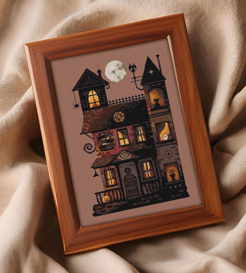 A framed cross-stitch image depicting a spooky, multi-level house with various architectural elements and a full moon in the sky.