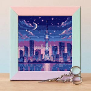 May include: A framed cross-stitch depicting a pixelated cityscape at night, with a prominent tower and a crescent moon. The frame is two-toned, with a pink inner border and a light green outer border. Decorative scissors rest in front.