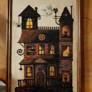 A framed cross-stitch image depicting a spooky, multi-story house with a full moon and a spider on the roof.