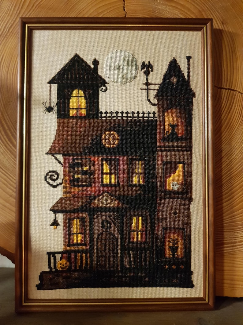 A framed cross-stitch image depicting a spooky, multi-story house with a full moon and a spider on the roof.