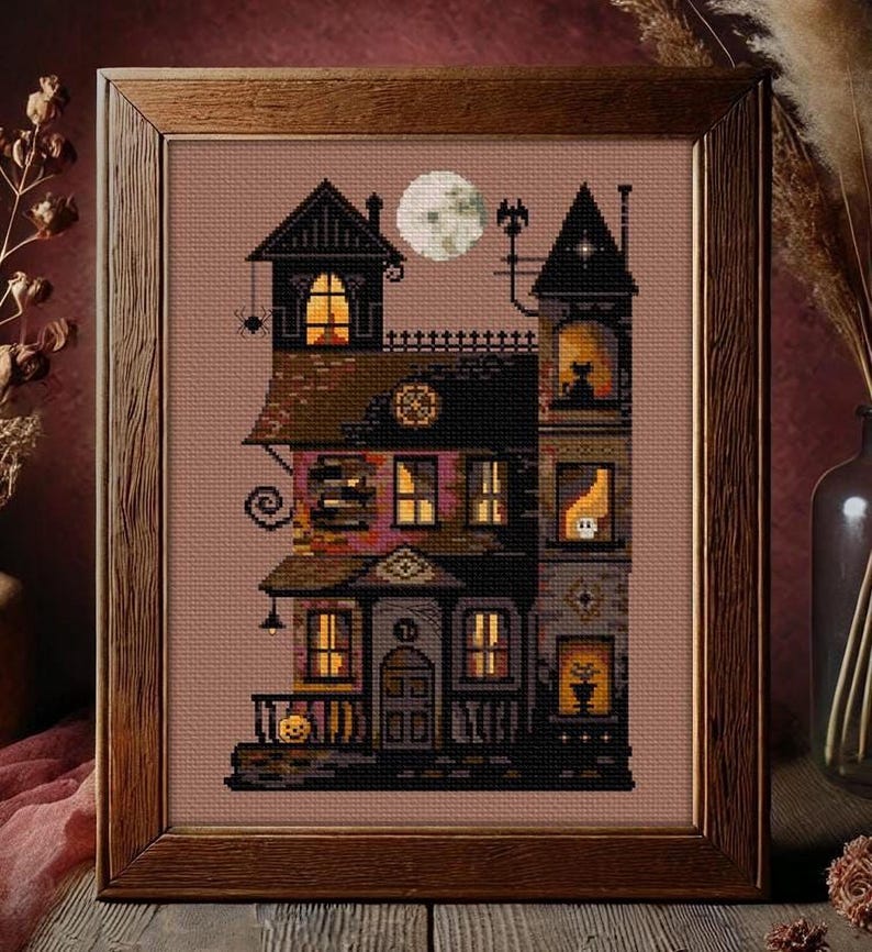 A framed cross-stitch image depicting a spooky, multi-story house with a full moon in the sky, surrounded by autumn foliage.