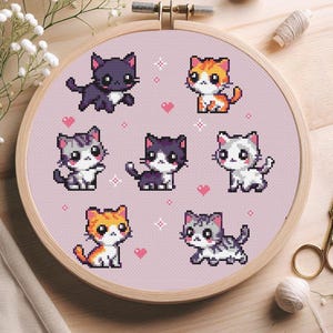 Könnte beinhalten: Ein Kreuzstich-Stickrahmen mit sieben pixeligen Katzendesigns in verschiedenen Farben, darunter Lila, Orange, Grau und Weiß, vor einem hellrosa Hintergrund. Das Design umfasst kleine rosa Herzen und Sternakzente.