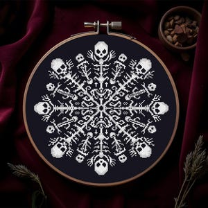 Könnte beinhalten: Ein Kreuzstich-Design eines Schneeflocks aus Totenköpfen und Knochen, präsentiert in einem hölzernen Stickrahmen. Das Design ist weiß auf einem dunkelblauen Hintergrund. Der Rahmen befindet sich vor einem dunkelroten Samthintergrund.