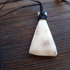 May include: A white bone pendant with a black braided cord. The pendant is shaped like a triangle with rounded edges and has a textured surface.